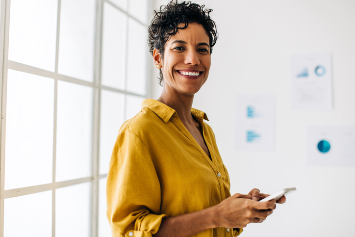 Happy female professional holding a mobile phone in her hand. Business woman browses business contacts on her phone as she stands next to a window in an office.